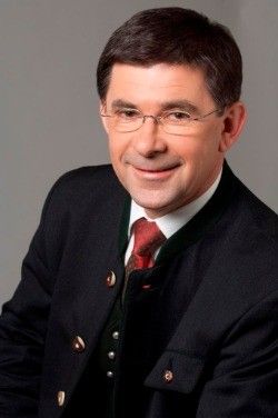 Ein Mann mit einer schwarzen Jacke, einem roten Krawatte und einer Brille lächelt und scheint für ein Foto zu posieren.