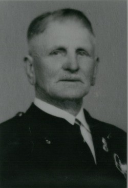 Ein Schwarz-Weiß-Porträt eines älteren Mannes in einem formellen Uniform, mit einem ernsten Ausdruck, vor einem schlichten Hintergrund stehend.