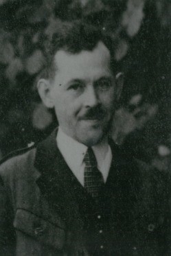 Ein altes Schwarz-Weiß-Foto eines Mannes in Uniform mit lächelndem Gesicht vor einem verschwommenen Hintergrund aus Blättern.