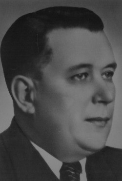 Eine Schwarz-Weiß-Fotografie eines Mannes mittleren Alters in formaler Kleidung, mit ernster Miene und kurzen, dunklen Haaren.