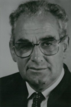 Schwarz-Weiß-Porträt eines Mannes mit Brille, Anzug und Krawatte. Er hat graues Haar und lächelt leicht. Der Hintergrund ist schlicht.