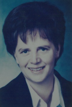 Eine Frau mit kurzen, gewellten Haaren und einem lächelnden Gesicht. Sie trägt einen schwarzen Blazer über einem weißen Hemd. Der Hintergrund ist hellblau.