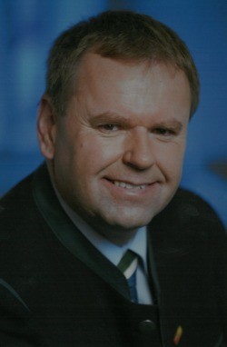 Ein lächelnder Mann in einem schwarzen Anzug und grüner Krawatte posiert für ein formelles Porträt vor blauem Hintergrund.