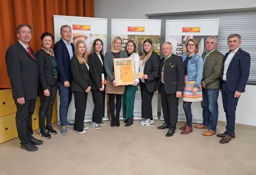 Eine Gruppe von Menschen steht gemeinsam für ein Foto. Eine Frau hält eine Urkunde. Hinter ihnen sind drei Banner ausgestellt, von denen eines Text und Bilder enthält.