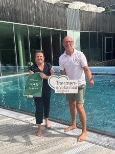 Ein Mann und eine Frau stehen barfuß in der Nähe eines Schwimmbeckens und halten grüne Schilder. Der Mann trägt ein weißes Hemd und Shorts, die Frau ein schwarzes Shirt und Hose. Hinter ihnen ist ein Gebäude mit Glaswänden zu sehen.