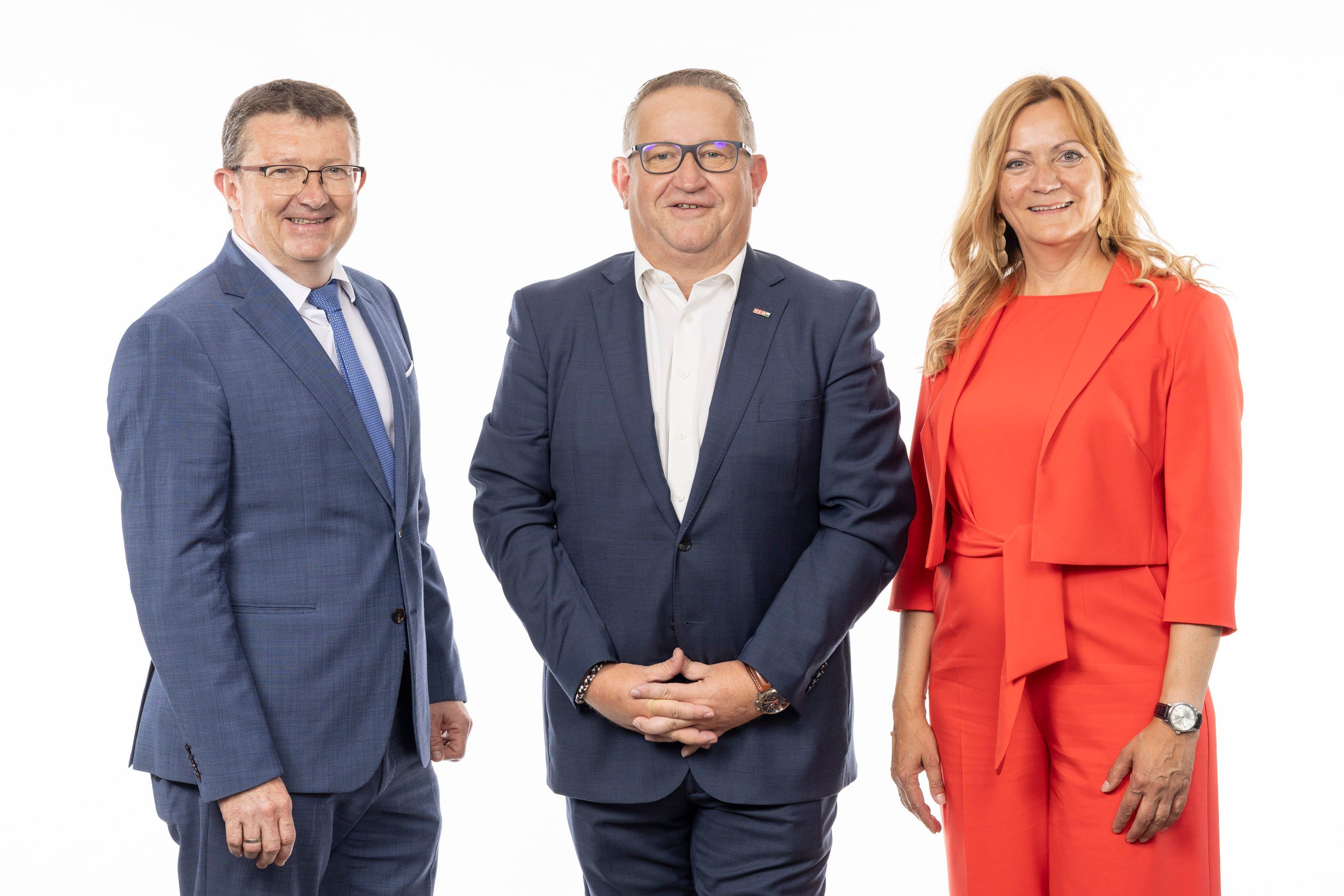 Drei Personen in formeller Kleidung stehen zusammen und lächeln für ein Foto. Der Mann links trägt einen dunkelblauen Anzug, der Mann in der Mitte einen marineblauen Anzug mit Brille und die Frau rechts ein leuchtend rotes Kleid.
