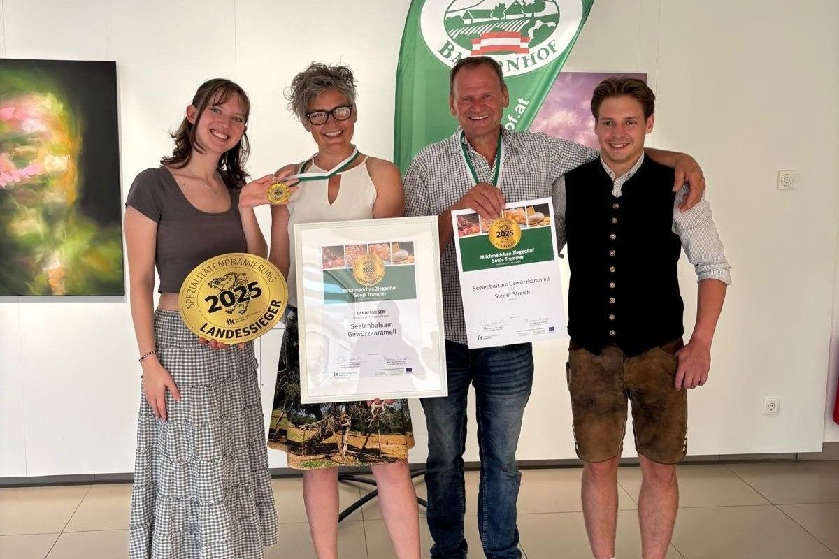 Vier Personen stehen lächelnd in einem Raum. Zwei Frauen halten Medaillen und Urkunden. Der Mann rechts trägt eine Weste und Shorts, während der Mann links ein kariertes Hemd trägt. Hinter ihnen ist eine grüne Fahne mit weißem Text.