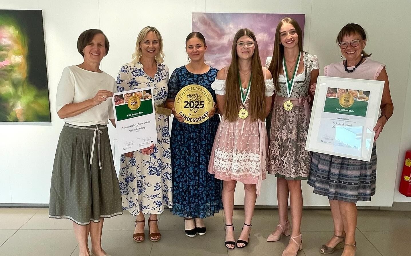 Fünf Frauen stehen zusammen und halten Auszeichnungen. Eine hält ein Zertifikat, eine andere einen goldenen Orden und drei tragen Medaillen. Sie stehen vor einer Kulisse mit einem Banner, das '2025 Landessiege' lautet.