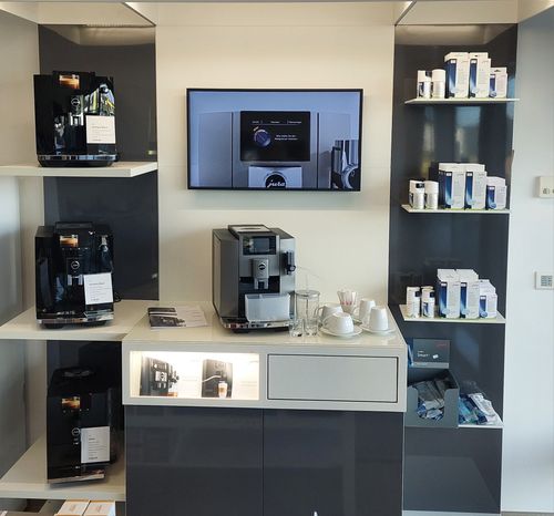 Ein moderner Küchenbereich mit Kaffeemaschine, Regalen und Fernseher. Die Kaffeemaschine steht auf dem Tresen, und es gibt Tassen, Untertassen und ein Glas.