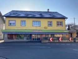 Ein gelbes Gebäude mit schwarzem Dach und grünem Vordach steht an der Straßenecke. Das Gebäude hat Glasfenster und Türen. Auf dem Bürgersteig befinden sich drei rote Schilder.