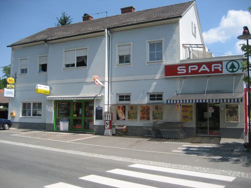 Außenansicht eines weißen Gebäudes mit einem roten Schild, das Spar steht. Es hat mehrere Fenster und eine grüne Tür. Es gibt Schilder und Poster an der Wand.