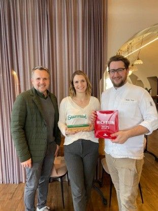 Drei Personen stehen drinnen, zwei Männer und eine Frau. Die Frau hält eine rote Box mit dem Wort Michelin darauf, während der Mann auf der rechten Seite ebenfalls eine ähnliche Box hält. Sie stehen vor einem Holzboden und einem lila Vorhang.