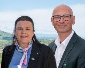 Ein Mann und eine Frau stehen lächelnd eng beieinander, mit einer wunderschönen Landschaft im Hintergrund. Der Mann trägt eine Brille und einen Anzug, während die Frau ein Tuch um den Hals hat.