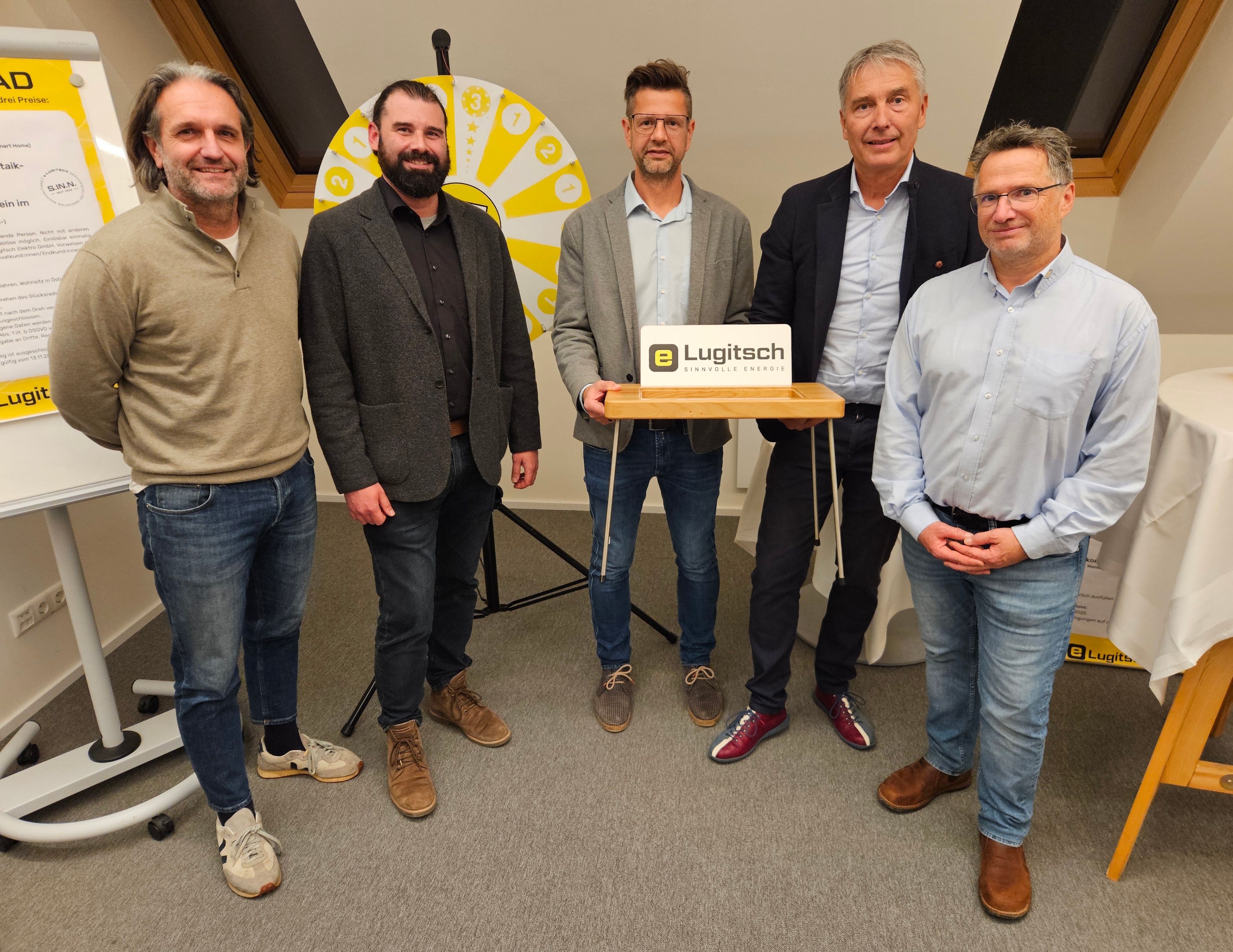 Fünf Männer stehen lächelnd vor einem Rad. Der Mann in der Mitte hält eine Box. Sie sind alle formell gekleidet. Hinter ihnen steht ein Stativ und eine Wand mit einem gelben Rad.