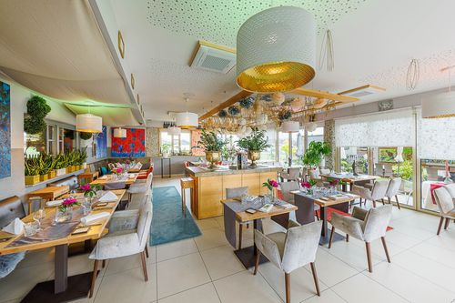 Ein modernes Restaurant mit großen Fenstern und einer warmen Atmosphäre. Tische sind mit Blumen und Grünpflanzen dekoriert, und eine große Deckenleuchte hängt über den Tischen.