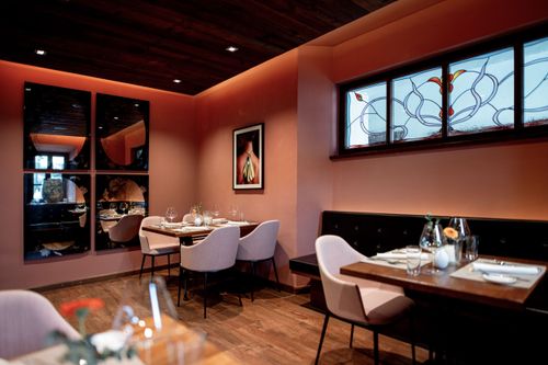 Ein kleines Restaurant mit modernem Innendesign, Holzböden, pfirsichfarbenen Wänden und einigen Tischen und Stühlen. An der Wand hängt ein gerahmtes Bild und ein Fenster mit Buntglas.