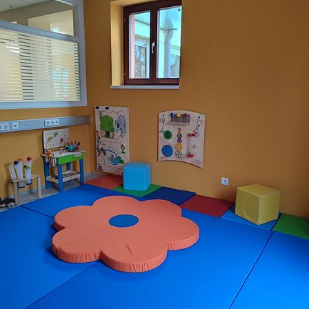 Ein Spielzimmer mit Blumenmatte, Würfeln und pädagogischen Postern. Es gibt ein Fenster mit Jalousien und ein kleiner Tisch mit Spielzeug.