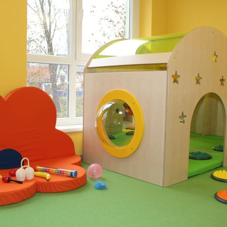 Ein Kinderspielbereich mit einem hölzernen Spielhaus, bunten Kissen, Spielzeug und einem Fenster, das einen Baum draußen zeigt.