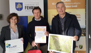 Drei Personen stehen vor einem Hintergrund mit den Texten 'Klar!' und 'Stadtgemeinde Fehring'. Sie halten ein gerahmtes Foto, ein Zertifikat und ein Stück Papier. Der Hintergrund hat auch ein Logo.