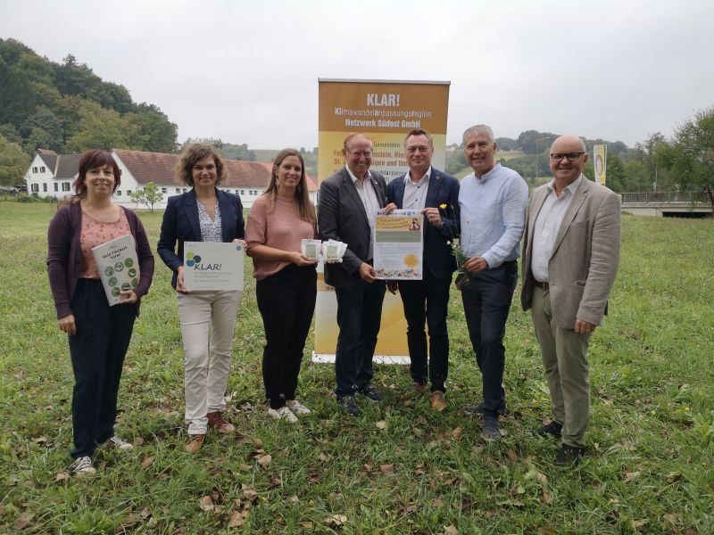 Eine Gruppe von Menschen steht auf einem Feld und hält Broschüren, Blumen und Papiere in den Händen. Sie lächeln und scheinen für ein Foto zu posieren. Hinter ihnen befindet sich ein gelbes Banner mit dem Text 'KLAR!' in weißen Buchstaben. In der Ferne befindet sich eine Brücke.