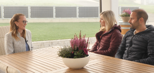 Drei Personen sitzen an einem Holztisch mit einer Pflanze. Eine Frau mit Brille und einer hellen Jacke, eine weitere Frau in einer dunklen Jacke und ein Mann in einer dunklen Jacke. Hinter ihnen ist ein großes Fenster mit Blick auf ein Feld.