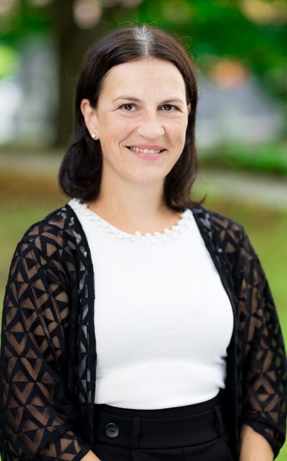 Eine Frau in einem weißen Oberteil und schwarzem Spitzencardigan, lächelt und steht im Freien in einem Park.