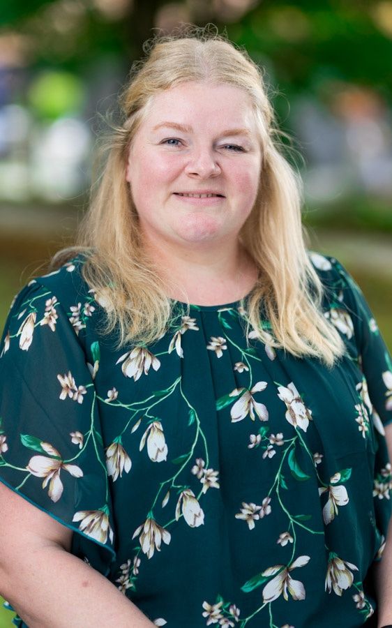Eine Frau mit blonden Haaren, die lächelt und ein grünes Blumenhemd trägt, steht in einem Park.