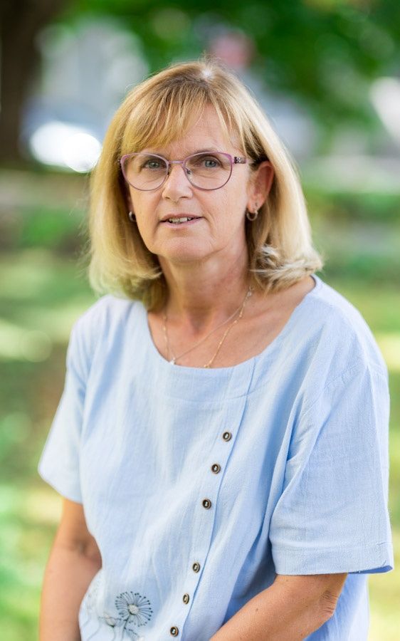 Eine Frau mit blonden Haaren, die eine blaue Bluse, eine Brille und eine Halskette trägt, steht in einem Park.