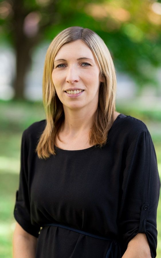 Eine Frau mit kurzen blonden Haaren und einem schwarzen Oberteil posiert für ein Bild in einem grünen Park.