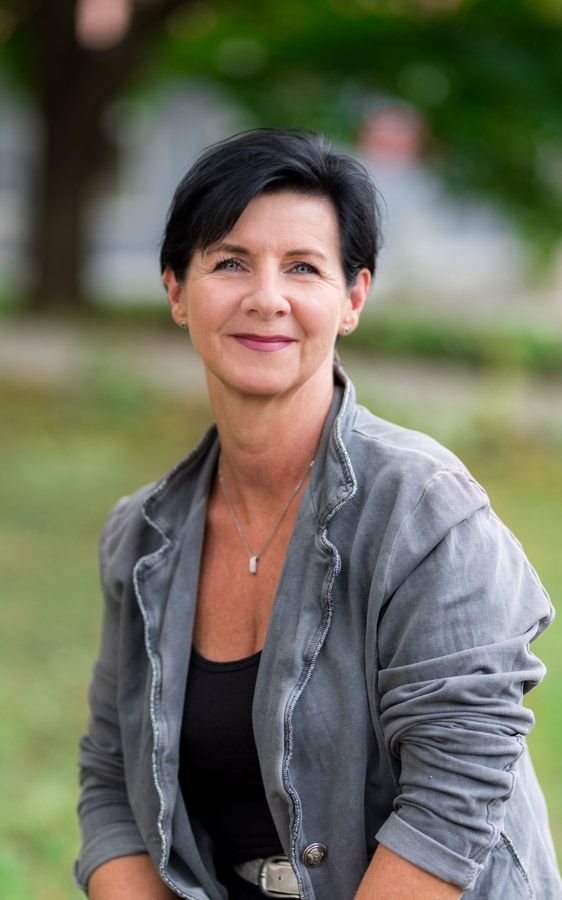 Eine Frau mit kurzen schwarzen Haaren, die eine graue Jacke und ein schwarzes Oberteil trägt, lächelt und steht auf einem grünen Rasen. Hinter ihr ein verschwommener Baum und ein grasiger Bereich.