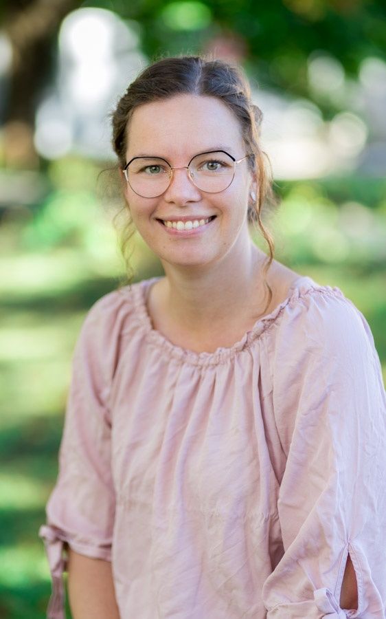 Eine Frau mit braunen Haaren, Brille und einem rosa Oberteil lächelt in die Kamera. Sie steht in einem Garten mit verschwommener Grünfläche im Hintergrund.