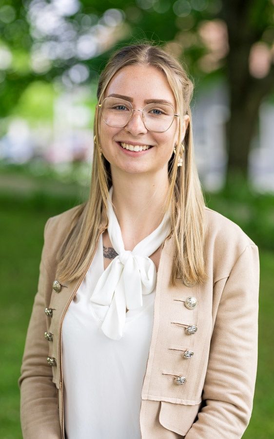 Eine lächelnde Frau mit blonden Haaren steht in einem Park. Sie trägt ein weißes Hemd, eine beige Jacke und eine Brille. Sie hat ein Tattoo am Hals.