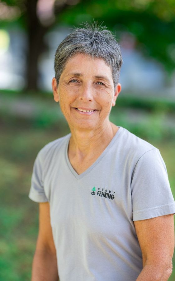 Eine ältere Frau mit grauem Haar lächelt und steht in einem Außenbereich. Sie trägt ein graues T-Shirt mit dem Stadt Fehring Logo. Der Hintergrund ist verschwommen mit grünen und weißen Farben.