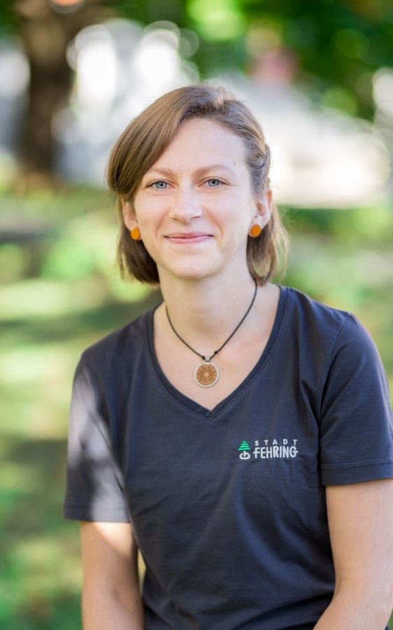 Eine lächelnde Frau mit kurzen Haaren, die ein dunkles V-Ausschnitt-Shirt mit Stadt Fehing trägt. Sie ist im Freien mit Bäumen im Hintergrund.