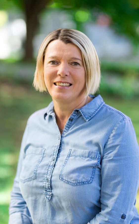 Eine Frau mit blondem Haar, lächelnd, trägt ein hellblaues Hemd mit Taschen und steht in einem Park.