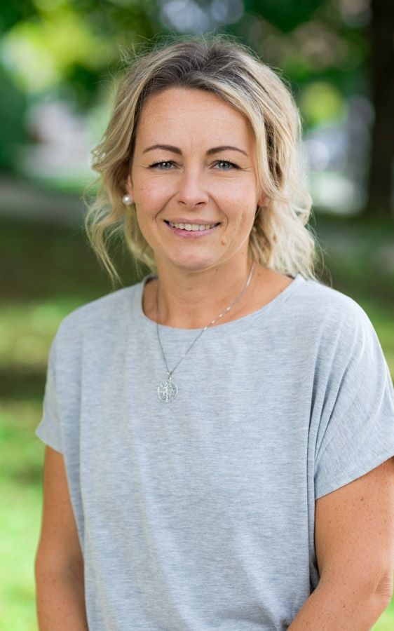 Eine blonde Frau mit einem Halsband lächelt in die Kamera im Park. Sie trägt ein graues T-Shirt.