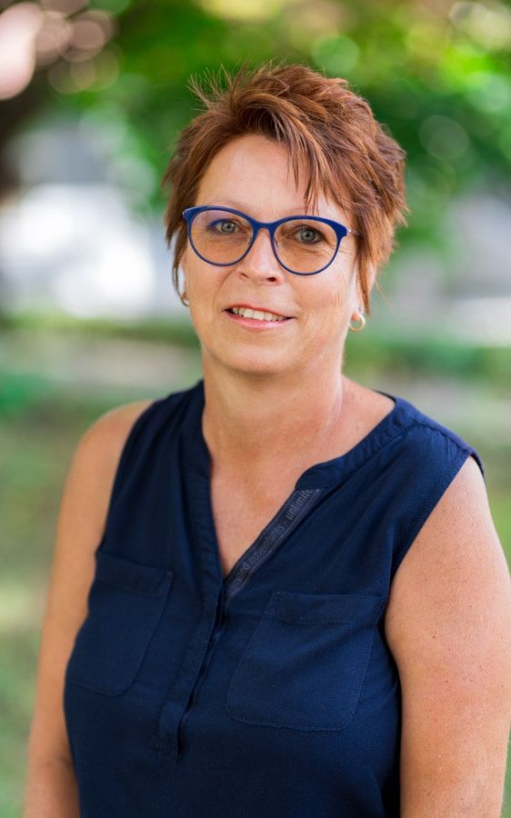 Eine Frau mit Brille und blauem Shirt posiert für ein Foto im Freien mit grünem Hintergrund.