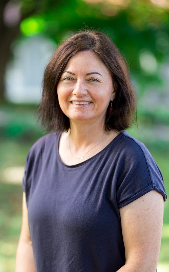 Eine Frau mit kurzen Haaren lächelt in die Kamera. Sie trägt ein blaues Shirt, Ohrringe und eine Halskette. Der Hintergrund ist unscharf.