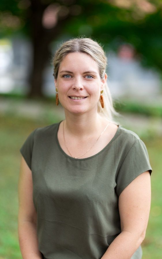Eine Frau mit blonden Haaren lächelt und trägt ein grünes Shirt und Ohrringe. Der Hintergrund ist unscharf mit Bäumen und Gras.
