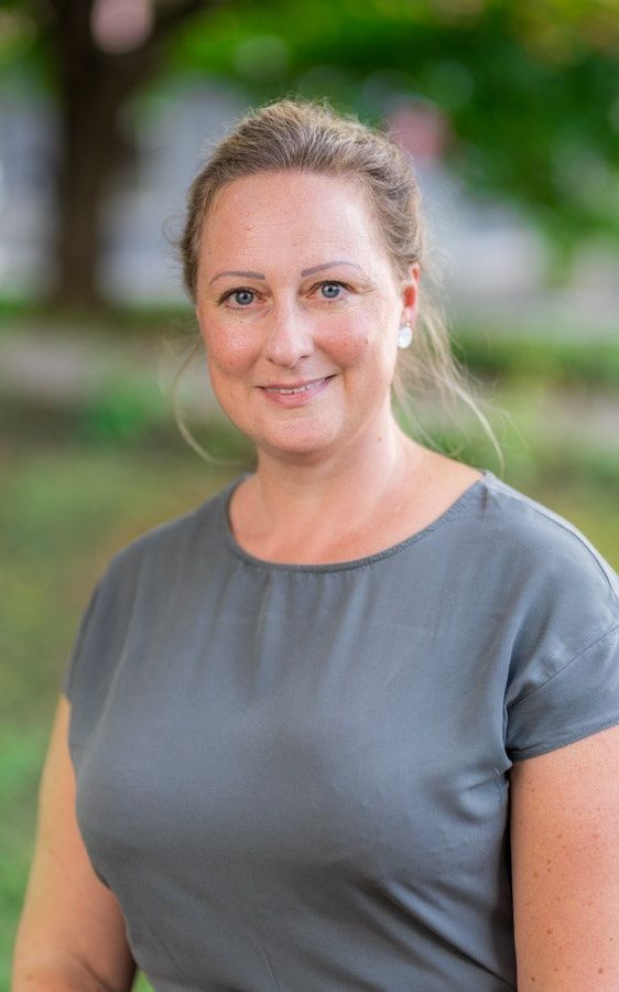Eine lächelnde Frau mit blonden Haaren steht in einem Park. Sie trägt ein graues Shirt und Ohrringe. Hinter ihr ist der Hintergrund unscharf mit grünem Gras und Bäumen.