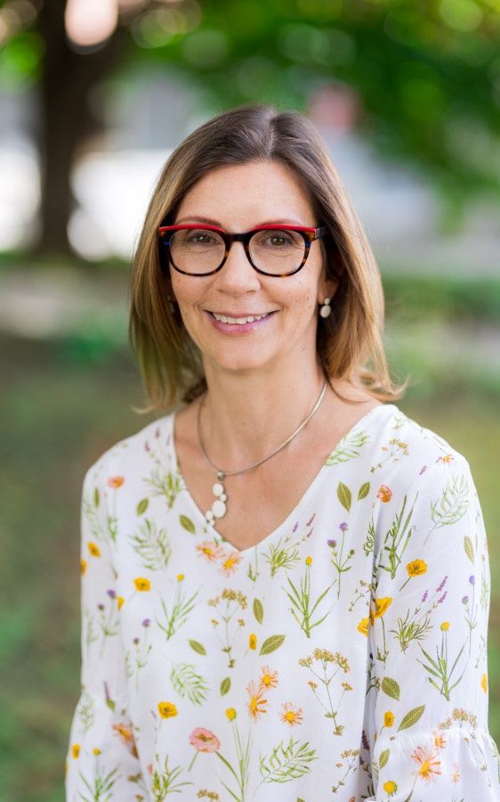 Eine lächelnde Frau in einem weißen Blumenblusen mit Brille und einer Halskette. Sie steht in einem Park mit verschwommener Grünfläche.