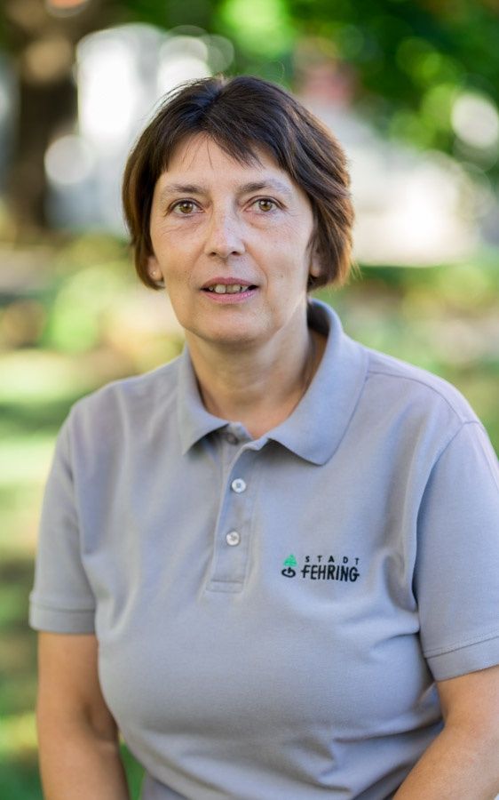 Eine Frau mit kurzen Haaren trägt ein hellblaues Polohemd mit den Worten Stadt Fehing darauf. Sie steht an einem Außenbereich.