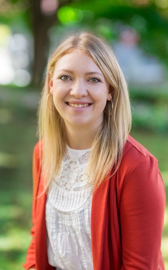 Eine lächelnde Frau mit blondem Haar steht im Freien. Sie trägt eine weiße Bluse und eine rote Jacke. Der Hintergrund ist unscharf, mit Bäumen und einem Auto.