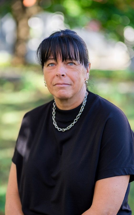 Eine Frau mit kurzen schwarzen Haaren trägt ein schwarzes Oberteil und eine silberne Kettenkette. Sie steht draußen.