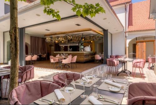 Ein elegantes Restaurant mit rosa Stühlen und Tischen, die für eine Mahlzeit vorbereitet sind. Der Tresen ist im Hintergrund zu sehen, mit Blick auf die Terrasse im Freien.