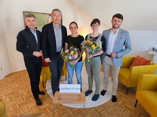 Fünf Personen stehen in einem Raum mit Holzboden, zwei Frauen mit Blumen, zwei Männer und eine Frau mit Brille. Sie lächeln und schauen zur Kamera. Ein Schild sagt 'wachsen & sein'.