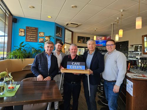 Fünf Männer stehen in einem Restaurant mit einem Schild, auf dem Café OZEan steht. Sie lächeln und scheinen für ein Foto zu posieren. Das Restaurant hat Holztische, blaue Wände und verschiedene Dekorationen.