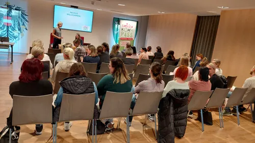 Ein Klassenzimmer mit Beamer zeigt eine Willkommensnachricht. Eine Frau steht vorne. Das Publikum sitzt aufmerksam, mit einem Banner hinter dem Sprecher, das Familien Komm Pass liest.