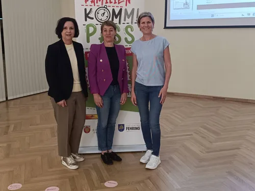 Drei Frauen stehen vor einem Banner mit einem Präsentationsbildschirm dahinter. Sie lächeln und scheinen für ein Foto zu posieren. Das Banner hat farbenfrohen Text und Logos.