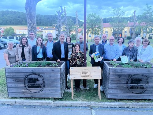 Eine Gruppe von Menschen steht um Holzpflanzgefäße herum und posiert für ein Foto. Sie halten Bücher in der Hand und scheinen an einer Gemeinschaftsgärtnerei-Veranstaltung teilzunehmen.
